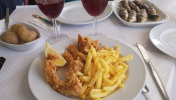 Fried fish, fries, and lemon slice on a plate with two drinks and boiled potatoes.