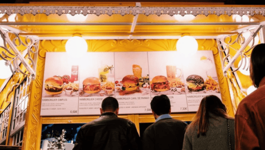 People standing at a food stall with lit menu displaying burgers.