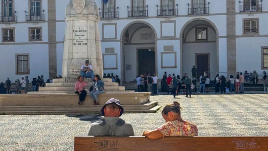 People sit on a bench in front of a large historic building and statue.