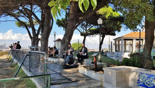 Musicians playing in a park with large trees and people sitting nearby.