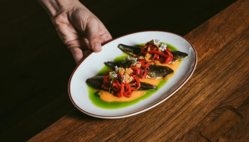 Hand placing a dish with sardines, red garnish, and green sauce on a wooden table.