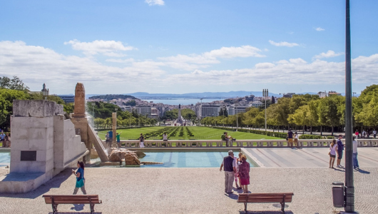 Park with wide green lawn, people walking, city and ocean in background.
