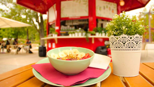 Fruit bowl on table near red food stand and plant.