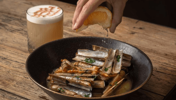 Hand squeezing lemon over clams in bowl, drink with foam on table.