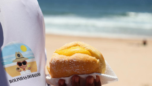 Person holding a pastry with cream filling, beach and ocean in background.