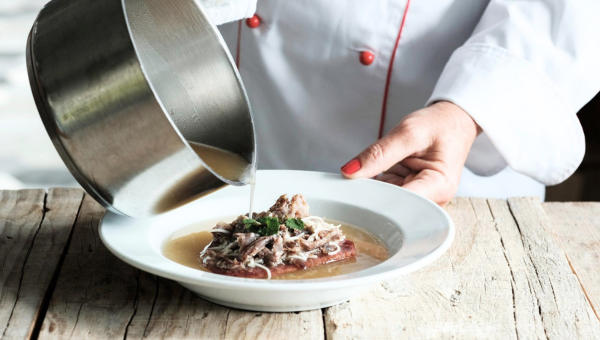 Chef pouring broth over shredded meat on a plate.