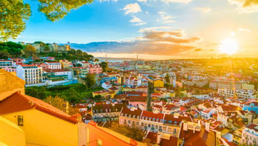Sunset view over a colorful, hilly cityscape with a castle in the distance and a vibrant sky.