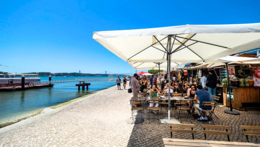 Outdoor café by a river with people sitting under large umbrellas on a sunny day.