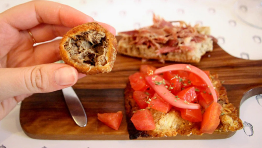 Hand holding bread, toasted slices with tomato and meat on a wooden board.
