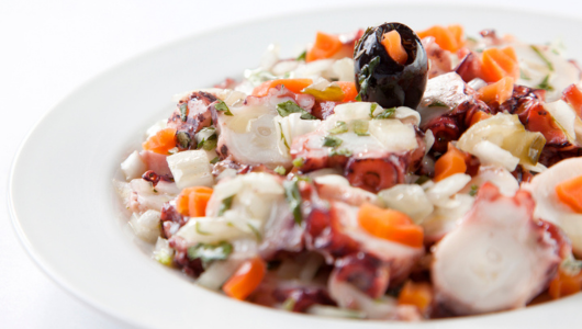 Seafood salad with octopus, vegetables, and black olive on a white plate.