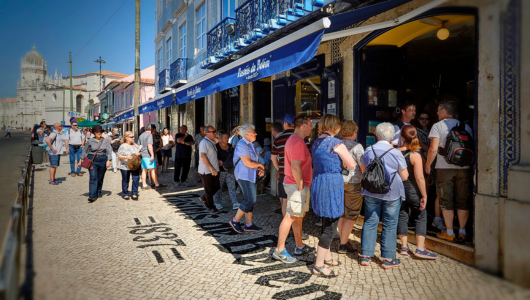 People in line outside a blue-fronted bakery on a sunny day.