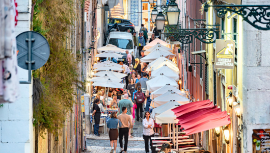 Narrow street with outdoor dining, umbrellas, and people walking.