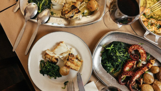 Plates of grilled seafood, greens, and potatoes with wine on a table.