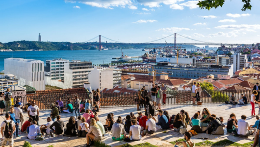 People sitting on a terrace overlooking a river and bridge in a cityscape.