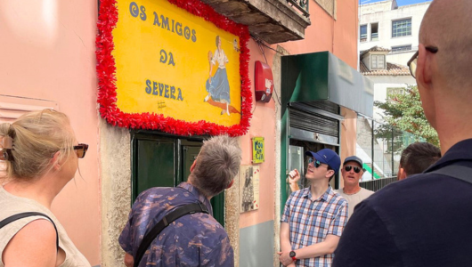 Group looking at a red-framed sign reading 'Os Amigos da Severa' on a city street.