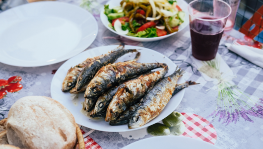 Grilled fish on a plate with salad, bread, and a glass of red drink on a table.
