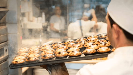 Baker holds tray of pastries in a bustling kitchen.