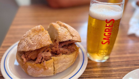 Sandwich with meat on a plate next to a glass of beer on a wooden table.