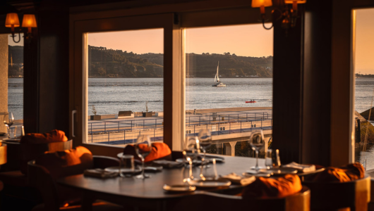 Cozy restaurant with view of sailboat on water through large windows.