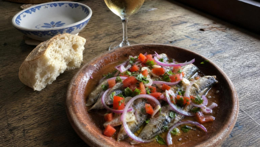 Plate of marinated sardines with onions and tomatoes, bread, and a glass of wine on a wooden table.