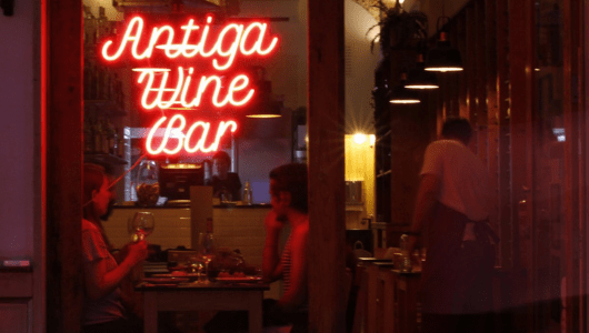 Neon sign 'Antiga Wine Bar' with people dining inside.