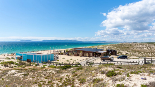Beachside building with cars, sandy dunes, and ocean under a blue sky with clouds.