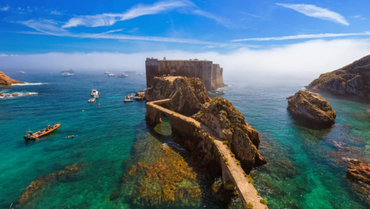 Stone path leads to island fort in clear blue sea with boats and clear sky.