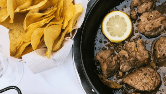 Meat stew with lemon slice in pan, chips in a bowl on white tablecloth.