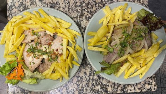 Two plates of meat with fries and salad on a speckled countertop.