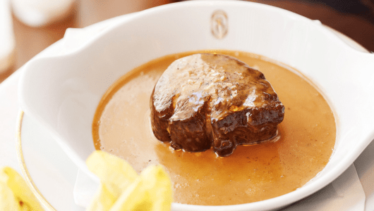 A piece of cooked meat in a rich brown sauce on a white plate.