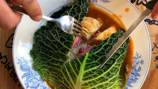 Hands cutting cabbage-wrapped dish with gravy on blue-patterned plate.