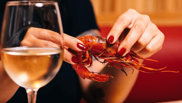 Person holding a cooked shrimp near a glass of white wine.