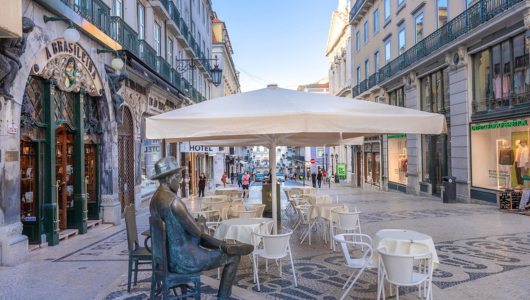 Outdoor cafe with statue and tables under an umbrella on a pedestrian street.