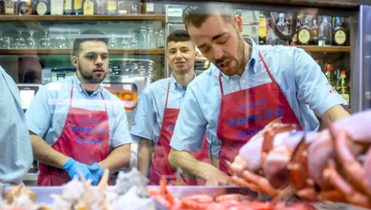 Men in uniforms prepare seafood at a counter in a market setting.