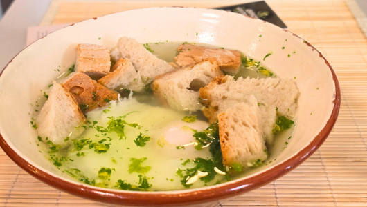 Soup with bread cubes, greens, and a poached egg in a ceramic bowl.