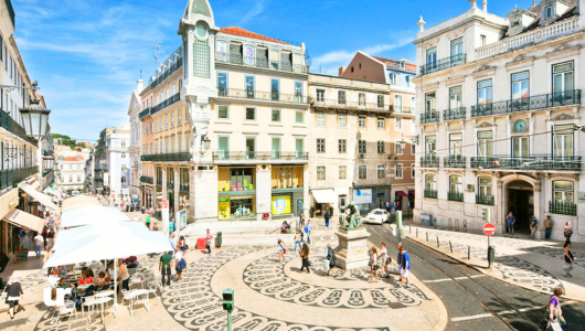 Sunny plaza with patterned paving, historic buildings, and people walking in Lisbon.