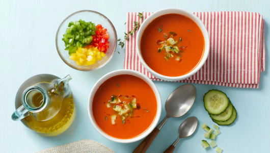 Two bowls of red soup with diced vegetables, oil bottle, and spoons on a striped cloth.