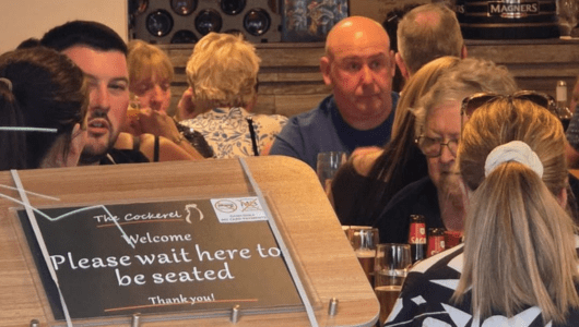 People dining in a restaurant, sign reads 'Please wait here to be seated'.