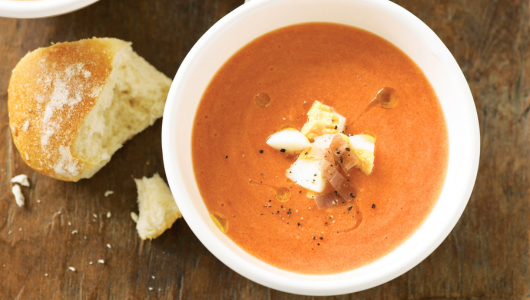 Bowl of tomato soup with croutons and pepper, next to a bread roll.