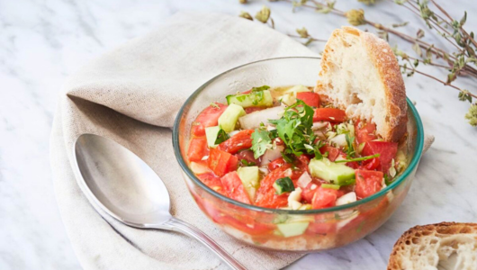 Bowl of gazpacho with bread slice on a napkin, spoon beside.