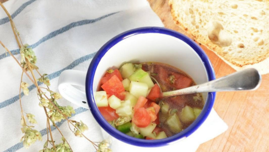 Cup of vegetable soup with spoon, bread slices, and flowers on a cloth.