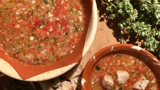 Two bowls of gazpacho with bread and herbs nearby on a wooden surface.