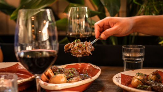 Hand holding fork with food over plates and wine glasses on a table.