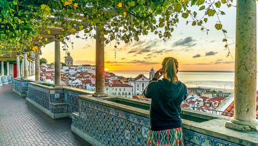Person photographing Lisbon cityscape at sunset from tiled balcony.