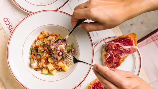 Person eating ceviche with fork, cured meat on toast beside.