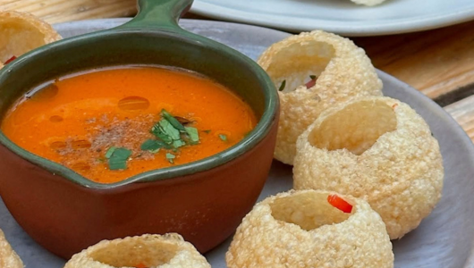 Pani puri with red sauce in a brown bowl on a plate.