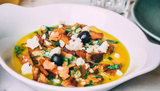 Yellow soup with croutons, cheese, and black olives in a white bowl.