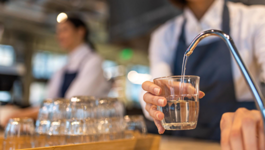 Person in apron filling glass with water from tap, blurred background.