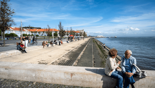 People sitting and walking along a waterfront promenade on a sunny day.