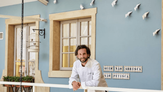 Man in white coat leaning on railing, blue wall with windows and bird decor behind him.